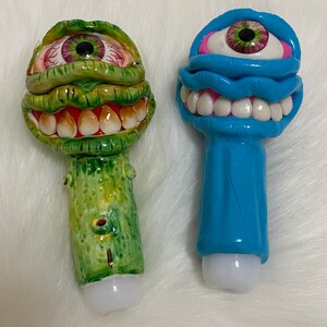May include: Two ceramic figurines with a whimsical, cartoonish style. One is green with a large eye and a toothy grin, the other is blue with a single eye and a wide, toothy smile.