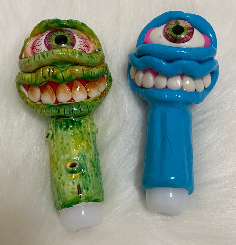May include: Two ceramic figurines with a whimsical, cartoonish style. One is green with a large eye and a toothy grin, the other is blue with a single eye and a wide, toothy smile.