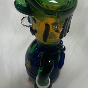 May include: A green glass figurine of a cartoon character with a green hat, blue overalls, and a white moustache. The figurine has a hole in the top of the hat for smoking.