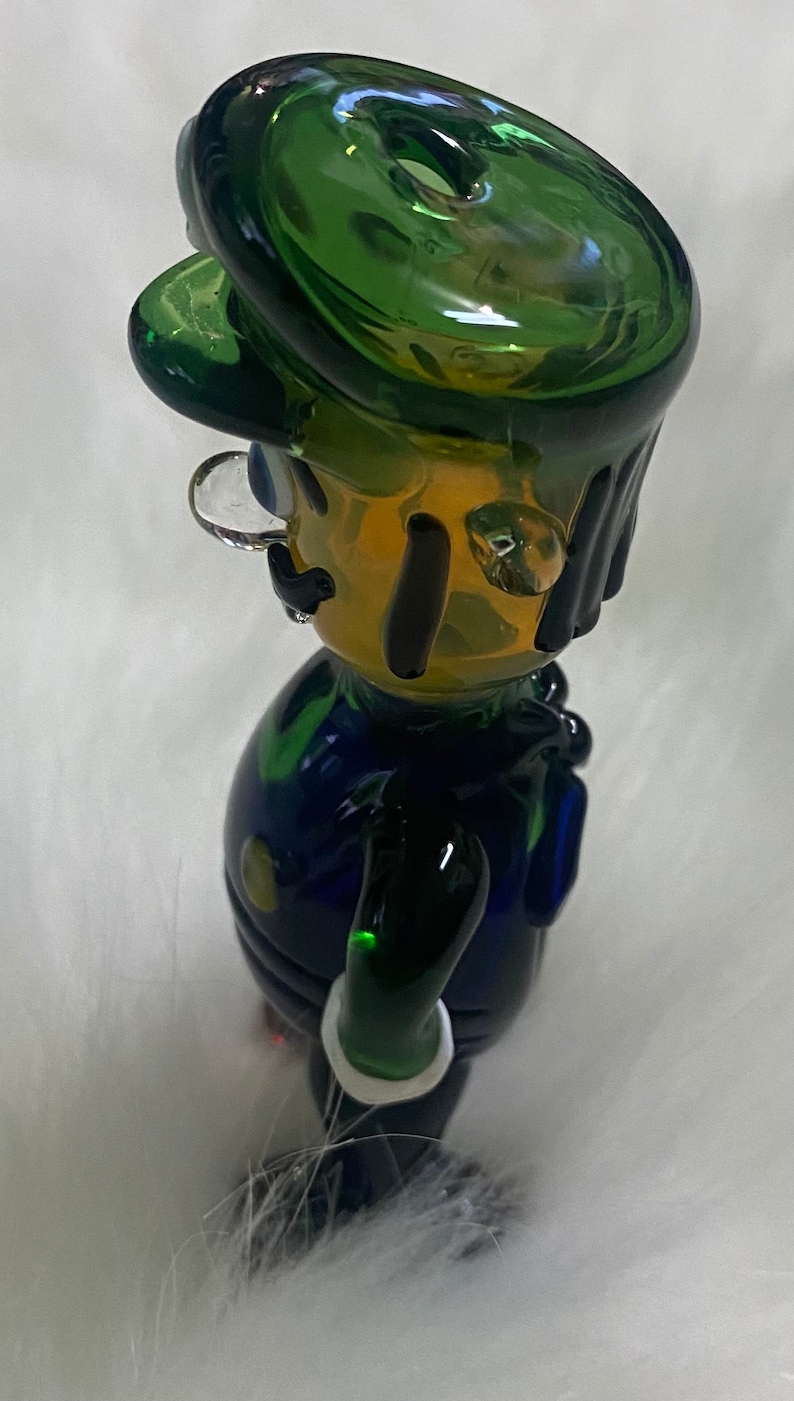 May include: A green glass figurine of a cartoon character with a green hat, blue overalls, and a white moustache. The figurine has a hole in the top of the hat for smoking.