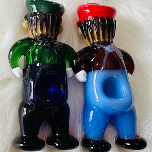 May include: Two glass figurines of people wearing overalls. The figurine on the left is wearing a green hat and has a blue bowl. The figurine on the right is wearing a red hat and has a light blue bowl.