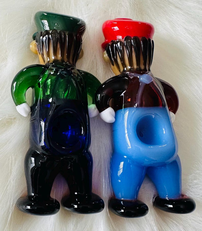 May include: Two glass figurines of people wearing overalls. The figurine on the left is wearing a green hat and has a blue bowl. The figurine on the right is wearing a red hat and has a light blue bowl.