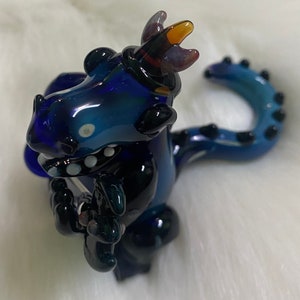 May include: A blue glass dragon figurine with a black tail and a purple and orange crown. The dragon is holding a small black object in its front paws.