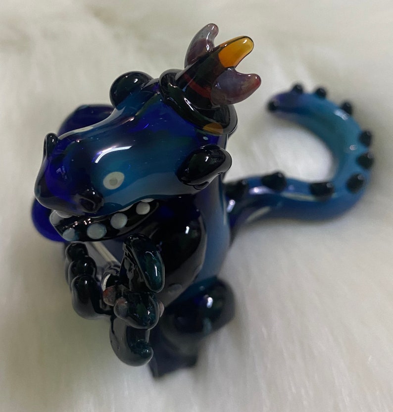 May include: A blue glass dragon figurine with a black tail and a purple and orange crown. The dragon is holding a small black object in its front paws.