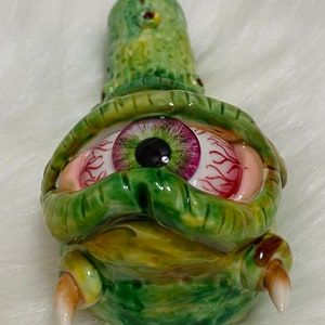 May include: Green ceramic pipe with a single large eye with a pink and green iris and red veins. The pipe has two small horns on the bottom.