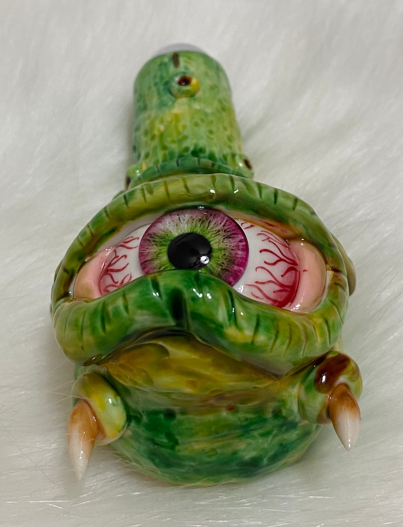 May include: Green ceramic pipe with a single large eye with a pink and green iris and red veins. The pipe has two small horns on the bottom.