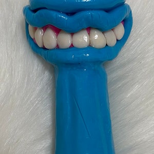 May include: A blue ceramic pipe with a single eye and a mouth with teeth. The eye is green and pink, and the mouth is pink with white teeth.