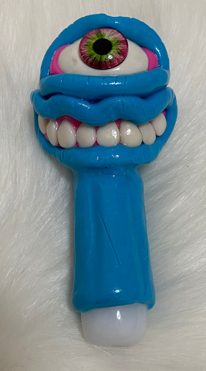 May include: A blue ceramic pipe with a single eye and a mouth with teeth. The eye is green and pink, and the mouth is pink with white teeth.