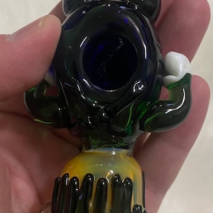 May include: A glass pipe with a black and green body, blue bowl, and white accents. The pipe is shaped like a cartoon character with arms and legs.