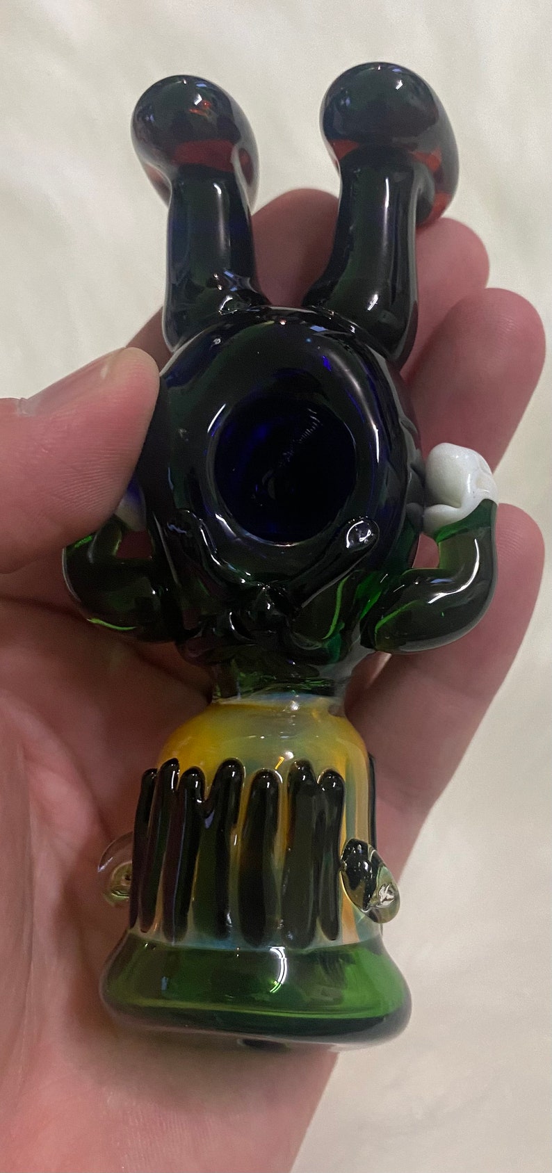 May include: A glass pipe with a black and green body, blue bowl, and white accents. The pipe is shaped like a cartoon character with arms and legs.