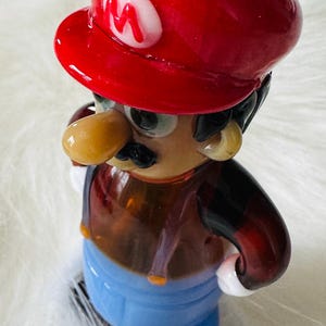 May include: A glass figurine of a cartoon character wearing a red hat with a white 'M' on it. The character has a brown nose, black moustache, and brown eyes. The figurine is wearing a brown shirt, blue trousers, and brown shoes.