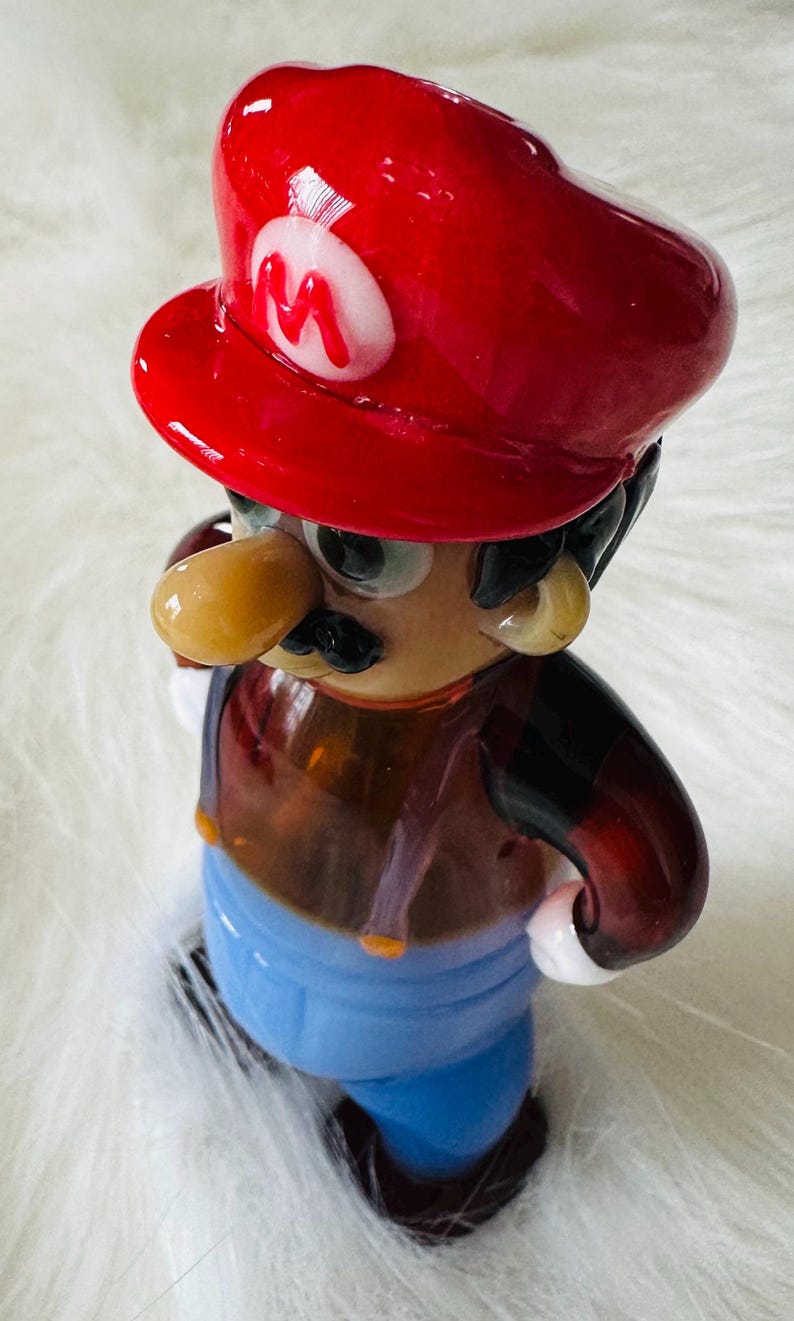 May include: A glass figurine of a cartoon character wearing a red hat with a white 'M' on it. The character has a brown nose, black moustache, and brown eyes. The figurine is wearing a brown shirt, blue trousers, and brown shoes.