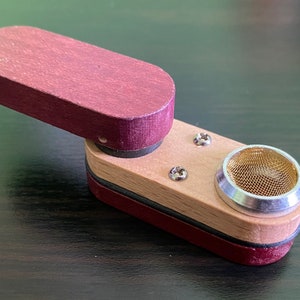 May include: A small, wooden pipe with a burgundy top and a natural wood base. The pipe has a metal bowl with a mesh screen.