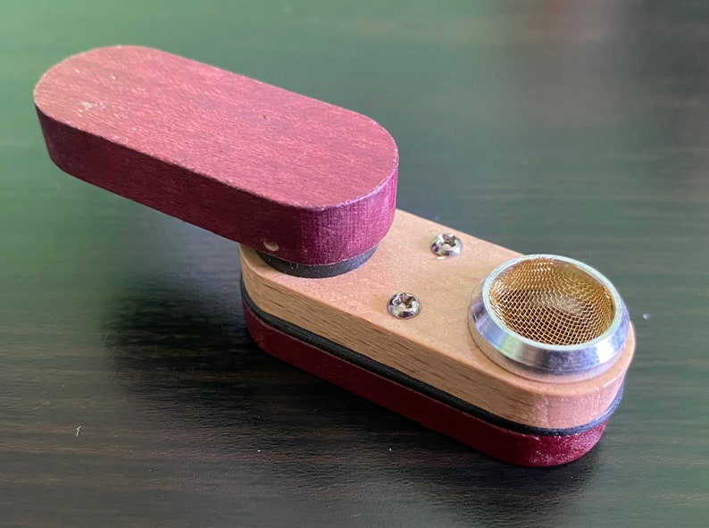 May include: A small, wooden pipe with a burgundy top and a natural wood base. The pipe has a metal bowl with a mesh screen.
