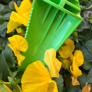 May include: A green plastic hand trowel with a serrated edge and a measurement scale marked in centimetres. The trowel is partially inserted into a bed of yellow pansies.
