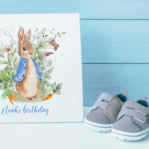 May include: A white card with a watercolor illustration of Peter Rabbit in a blue jacket surrounded by green leaves and flowers. The text "Noah's birthday" is printed in purple at the bottom of the card.