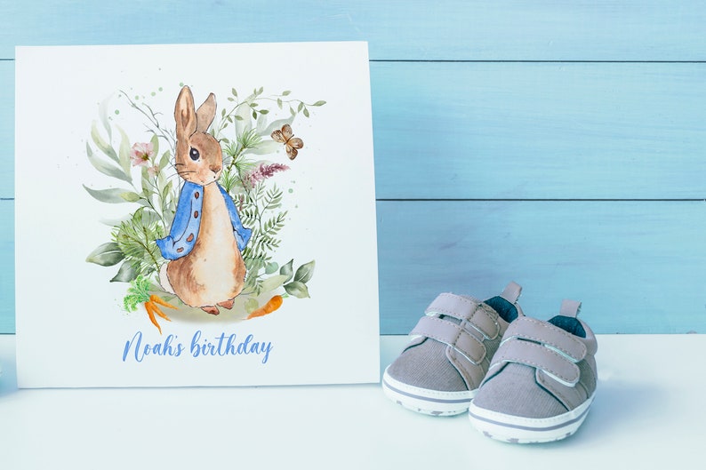 May include: A white card with a watercolor illustration of Peter Rabbit in a blue jacket surrounded by green leaves and flowers. The text "Noah's birthday" is printed in purple at the bottom of the card.