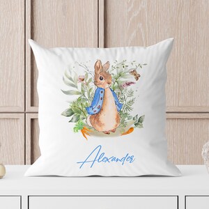 May include: White pillow with a watercolor illustration of a rabbit wearing a blue jacket and surrounded by green foliage. The name "Alexander" is written in blue cursive below the rabbit.