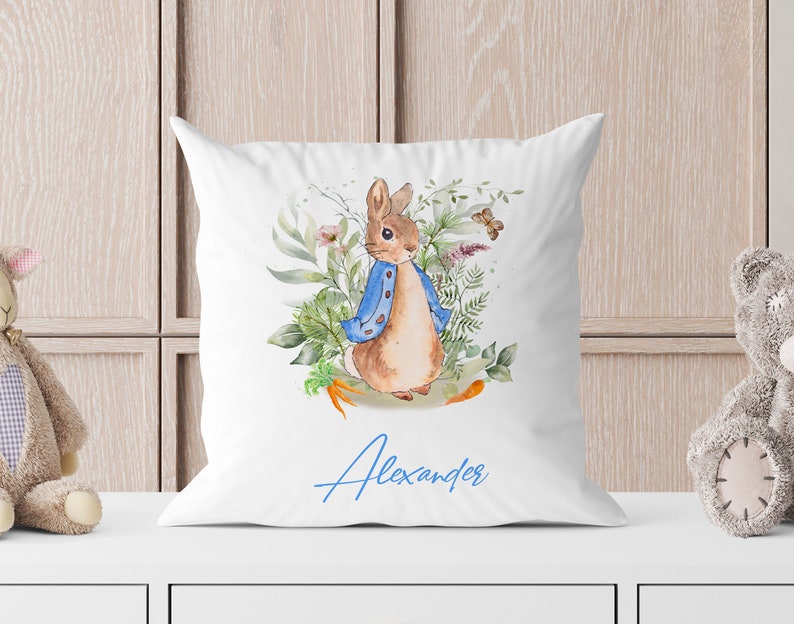 May include: White pillow with a watercolor illustration of a rabbit wearing a blue jacket and surrounded by green foliage. The name "Alexander" is written in blue cursive below the rabbit.