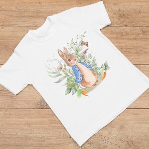 May include: White t-shirt with a watercolor illustration of Peter Rabbit in a blue jacket, surrounded by green leaves and flowers.
