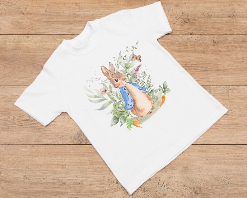 May include: White t-shirt with a watercolor illustration of Peter Rabbit in a blue jacket, surrounded by green leaves and flowers.