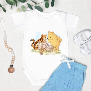 K&ouml;nnte beinhalten: Wei&szlig;er Baby-Strampler mit einer Illustration von Winnie Puuh, Ferkel und Tiger, die ein Picknick machen. Der Strampler wird mit einer hellblauen Babyhose und einem Holz-Bei&szlig;ring kombiniert.