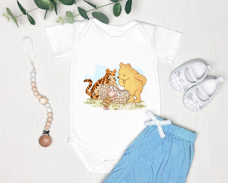 K&ouml;nnte beinhalten: Wei&szlig;er Baby-Strampler mit einer Illustration von Winnie Puuh, Ferkel und Tiger, die ein Picknick machen. Der Strampler wird mit einer hellblauen Babyhose und einem Holz-Bei&szlig;ring kombiniert.