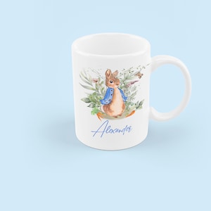 May include: White ceramic mug with a watercolor illustration of Peter Rabbit in a blue jacket surrounded by green foliage. The name "Alexander" is written in blue cursive at the bottom of the mug.