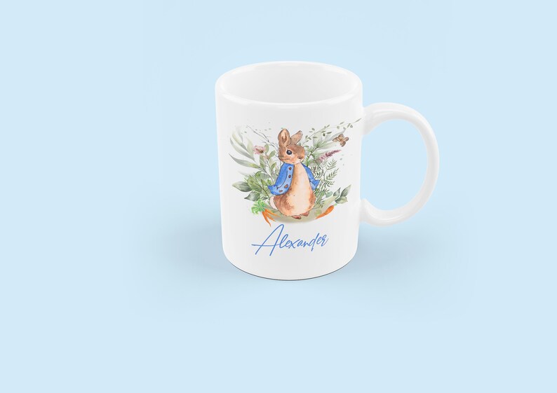 May include: White ceramic mug with a watercolor illustration of Peter Rabbit in a blue jacket surrounded by green foliage. The name "Alexander" is written in blue cursive at the bottom of the mug.