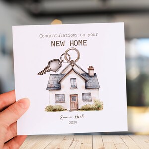 May include: A white card with a watercolor illustration of a house with a key on a ring. The card says "Congratulations on your NEW HOME" and "Emma & Noah 2024".