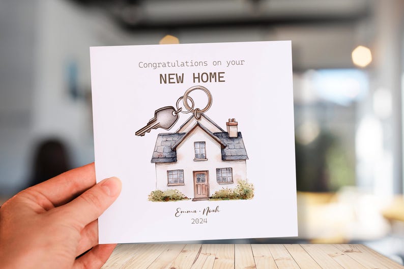 May include: A white card with a watercolor illustration of a house with a key on a ring. The card says "Congratulations on your NEW HOME" and "Emma & Noah 2024".