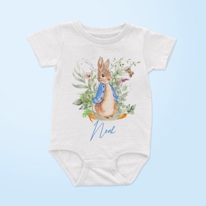May include: White baby onesie with a watercolor illustration of Peter Rabbit in a blue jacket surrounded by green foliage and a butterfly. The onesie has the name "Noah" written in blue cursive.