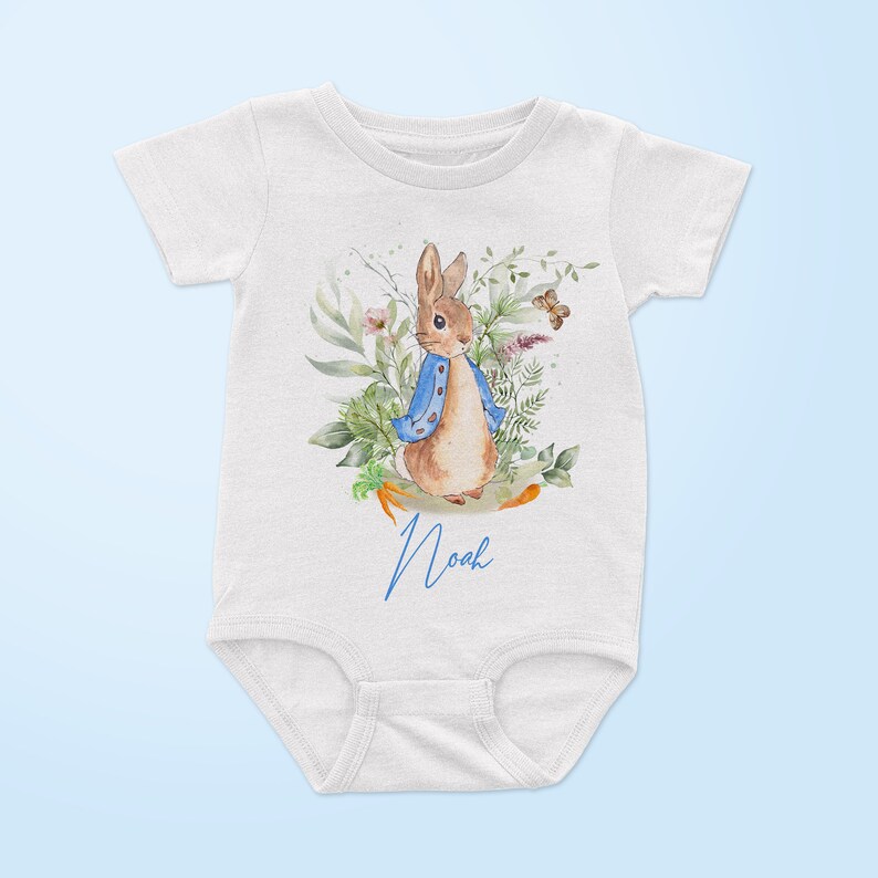 May include: White baby onesie with a watercolor illustration of Peter Rabbit in a blue jacket surrounded by green foliage and a butterfly. The onesie has the name "Noah" written in blue cursive.
