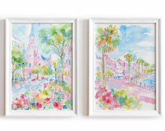 May include: Two framed watercolor prints. One depicts a pink church and street scene with colorful flowers. The other shows a waterfront with palm trees and buildings, also in watercolor. Both are in white frames.