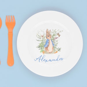 May include: A white ceramic plate with a watercolor illustration of Peter Rabbit in a blue jacket surrounded by green foliage. The plate has the name "Alexander" written in blue cursive.