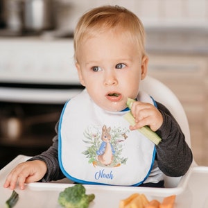 May include: A white baby bib with a blue trim featuring a watercolor illustration of Peter Rabbit with the name "Noah" embroidered in blue.