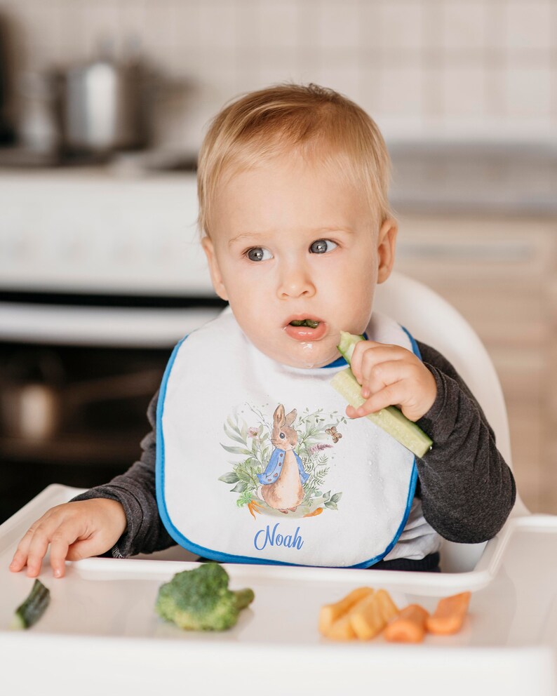 May include: A white baby bib with a blue trim featuring a watercolor illustration of Peter Rabbit with the name "Noah" embroidered in blue.