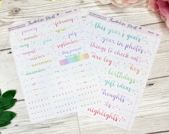 Op de afbeelding: Twee vellen met kleurrijke planner stickers met regenboog letters. Het eerste vel heeft de maanden van het jaar en het tweede vel heeft zinnen zoals "de doelen voor dit jaar" en "cadeau-ideeën".
