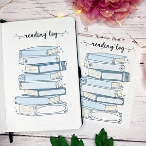 Pode incluir: Um diário de leitura e uma folha de adesivos com uma pilha de livros azul claro. O diário está aberto, revelando páginas pontilhadas. A folha de adesivos tem um design semelhante. As palavras "reading log" estão escritas em itálico.