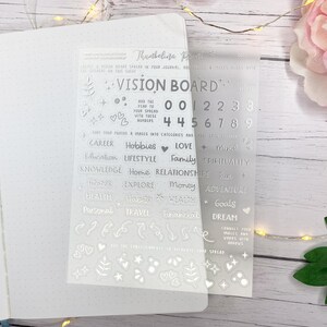 Op de afbeelding: Transparante vision board stickers met tekst en cijfers, waaronder "Vision Board", "Career", "Hobbies" en de cijfers 0-9. De stickers zitten op een transparante folie, naast een open notitieboek.