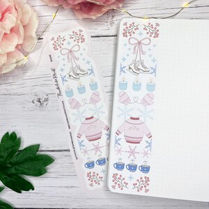 May include: A winter-themed bookmark and notebook with matching designs. The bookmark and notebook border feature ice skates, candles, mittens, sweaters, snowflakes, and teacups in pink, blue, and white. The bookmark has the text "Theodelite Print".