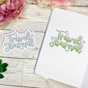 May include: A white notebook with a dotted pattern and the text "Travel Journal" written in green and black. A sticker with the same text is on a wooden surface.