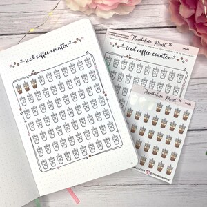Könnte beinhalten: Ein weißes Notizbuch und Aufkleberbögen mit Illustrationen von Eiskaffeebechern. Das Notizbuch hat ein gepunktetes Raster und den Text "iced coffee counter". Die Aufkleberbögen tragen den Text "Thumbelina Print".