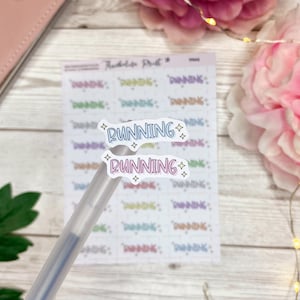 Op de afbeelding: Twee witte stickers met het woord "RUNNING" in blauwe en roze letters met glitteraccenten. De stickers bevinden zich op een vel met transparante stickers met een verscheidenheid aan kleurrijke woorden.