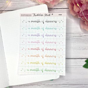 May include: A sticker sheet with the text "a month of dinners" in a rainbow gradient font. The text is repeated multiple times on a grid background. The sticker sheet is on top of an open notebook.