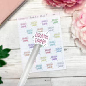 May include: A sheet of stickers with the words "Brain Dump" in pink, with star accents. The stickers are on a white background with a grid pattern. A clear pen is in the foreground, with a pink wallet and flowers in the background.
