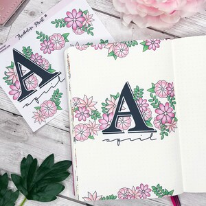 May include: A sticker sheet with a floral border and the letter A with the word "april" written below it. The sticker sheet is on a white wooden surface.