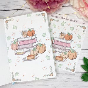 Könnte beinhalten: Ein weißes Notizbuch und ein Stickerbogen mit Herbstmotiven. Das Design zeigt Bücherstapel, Kürbisse, Teetassen und Kuchenstücke. Das Notizbuch ist geöffnet, und der Stickerbogen liegt daneben.