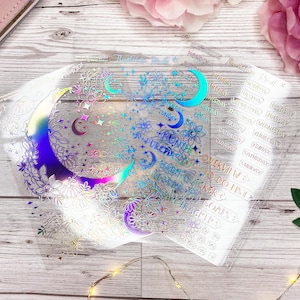 Könnte beinhalten: Ein Satz transparenter, schimmernder Planer-Aufkleber mit Mond- und Blumenmotiv. Die Aufkleber enthalten die Wochentage und die Monate des Jahres.