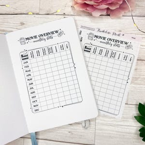 May include: An open journal and a separate paper sheet featuring a black and white printed movie overview monthly stats tracker. The grid includes columns for movies watched, bought, watched at home, in the cinema, genres, 5 star movies, and DNF.
